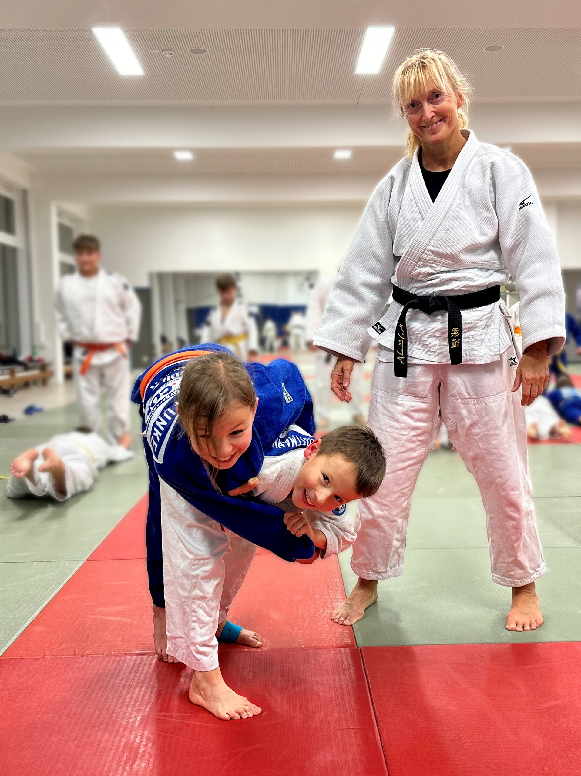 Andrea Goslar lächelt im weißen Judo-Anzug mit schwarzem Gürtel in einer Trainingshalle. Im Vordergrund zwei Kinder im Judo-Anzug, die kämpfen und lächeln.