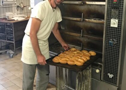 Lächelnder Bäcker in weißer Arbeitskleidung zieht ein Blech frisch gebackener Brötchen aus dem großen Backofen in der Backstube.