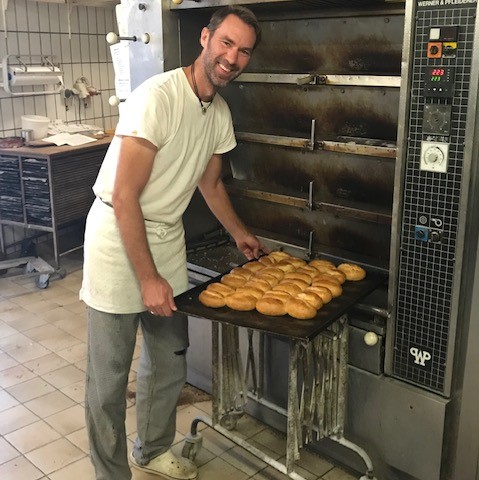 Lächelnder Bäcker in weißer Arbeitskleidung zieht ein Blech frisch gebackener Brötchen aus dem großen Backofen in der Backstube.