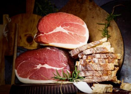 Zwei große Schinkenstücke übereinander gestapelt, daneben aufgeschnittenes Landbrot und ein Rosmarinzweig auf Holzuntergrund.