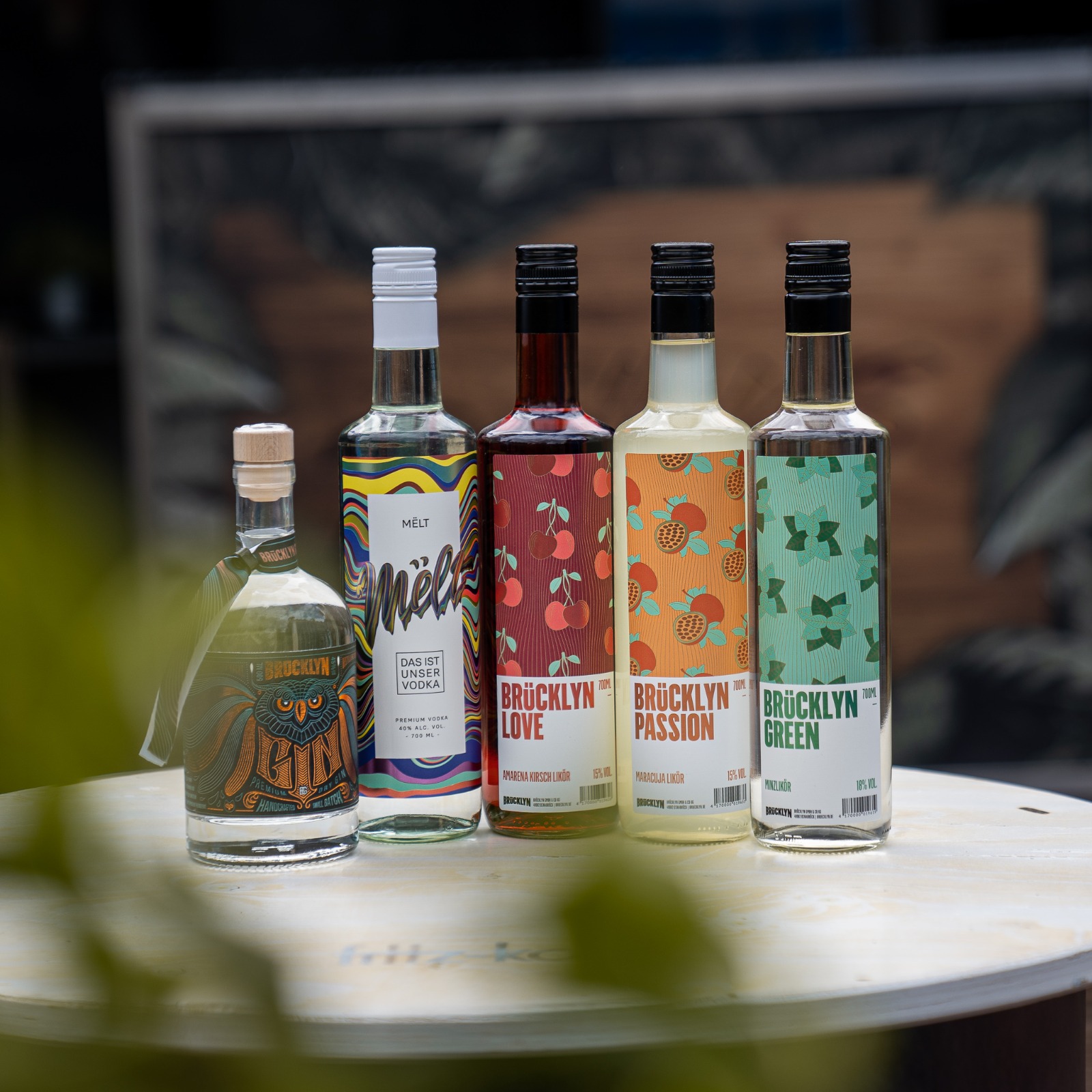 Fünf Flaschen Spirituosen nebeneinander auf einem Tisch, darunter Gin, Wodka und Liköre mit folgendem Schriftzug: Brücklyn und Mēlt.