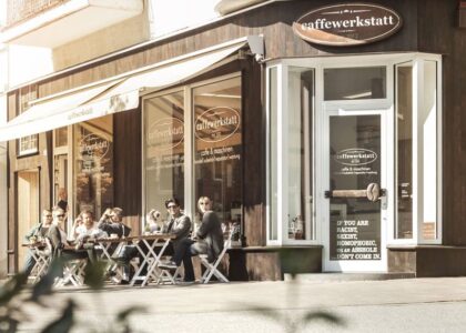 Gäste sitzen bei sonnigem Wetter vor dem Café caffewerkstatt an Tischen im Freien unter einer Markise.
