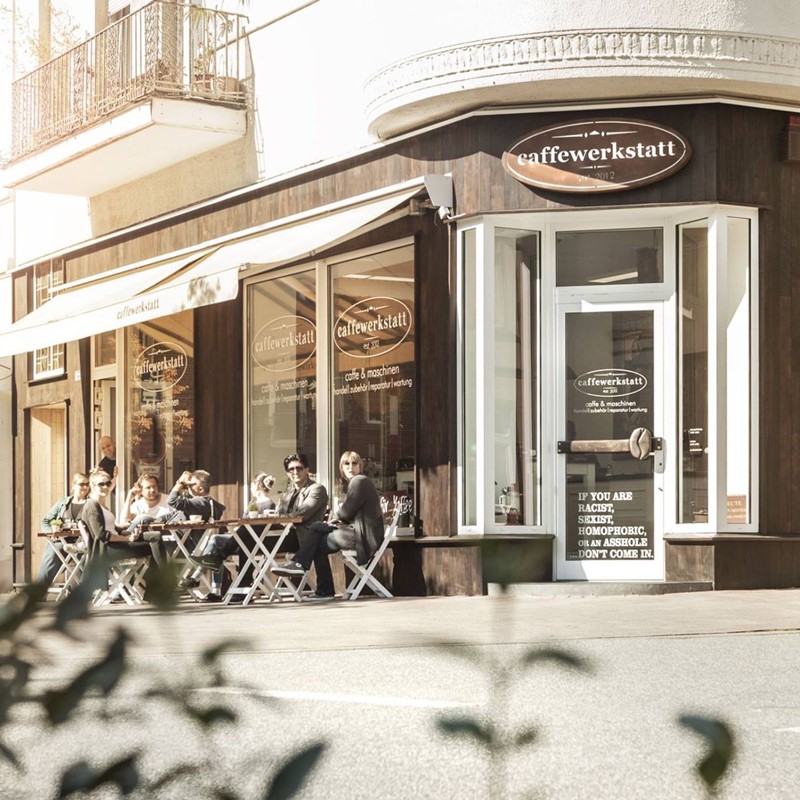 Gäste sitzen bei sonnigem Wetter vor dem Café caffewerkstatt an Tischen im Freien unter einer Markise.