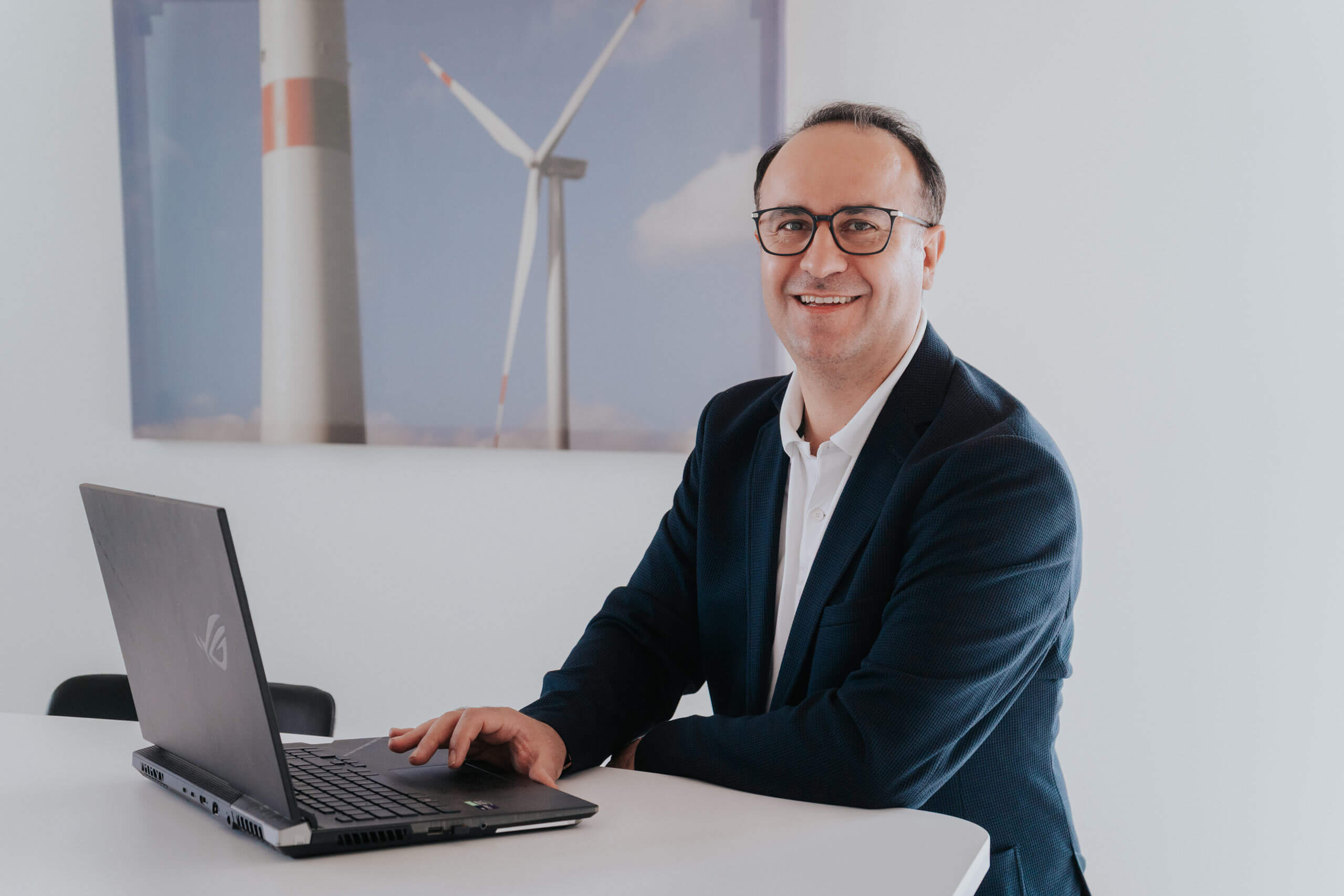 Lächelnder Mann mit Brille im Sakko sitzt am Tisch vor einem Laptop; im Hintergrund ein Windrad-Poster.