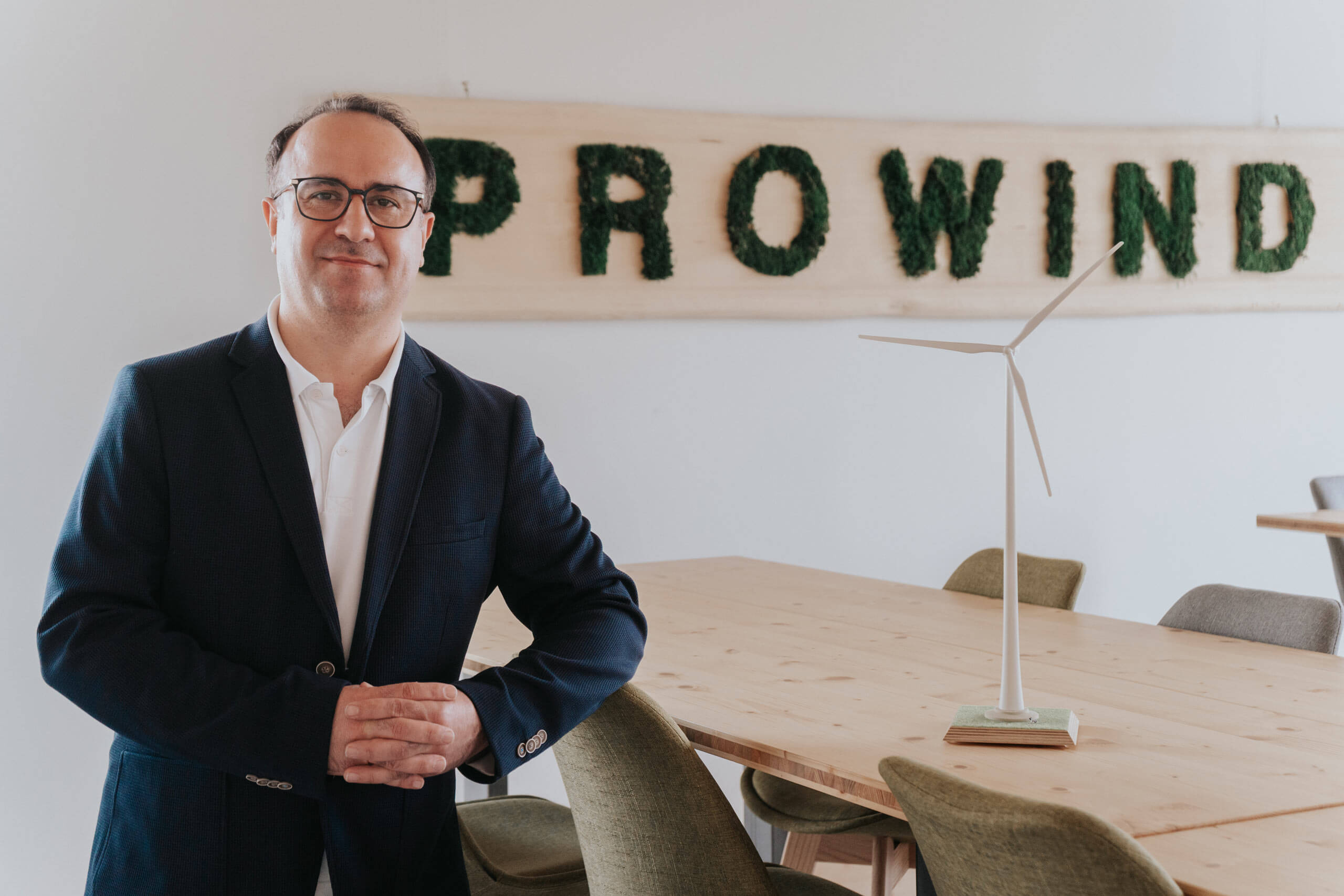 Mann im Sakko steht im Büro neben einem Tisch mit Windrad-Modell und dem Schriftzug „PROWIND“ an der Wand.