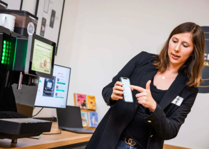 Eine Frau im dunklen Blazer zeigt auf ihr Smartphone neben einer modernen Kaffeemaschine im Büro.