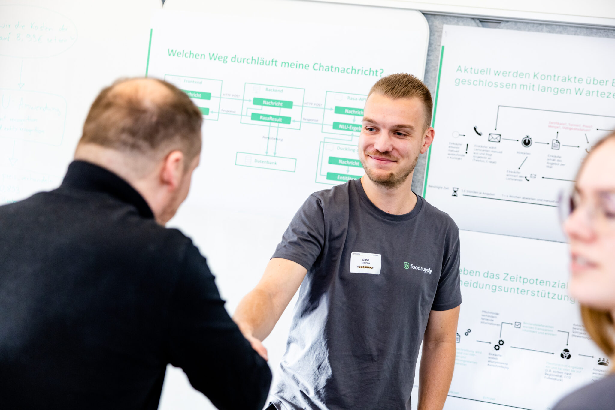 Ein lächelnder junger Mann im grauen T-Shirt gibt einer Person vor einer Präsentationswand den Handschlag.
