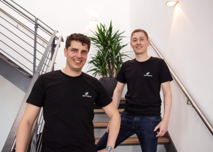 Zwei lächelnde Männer im schwarzen T-Shirt der G&S IT Group stehen gemeinsam auf einer modernen Holztreppe im Büro.