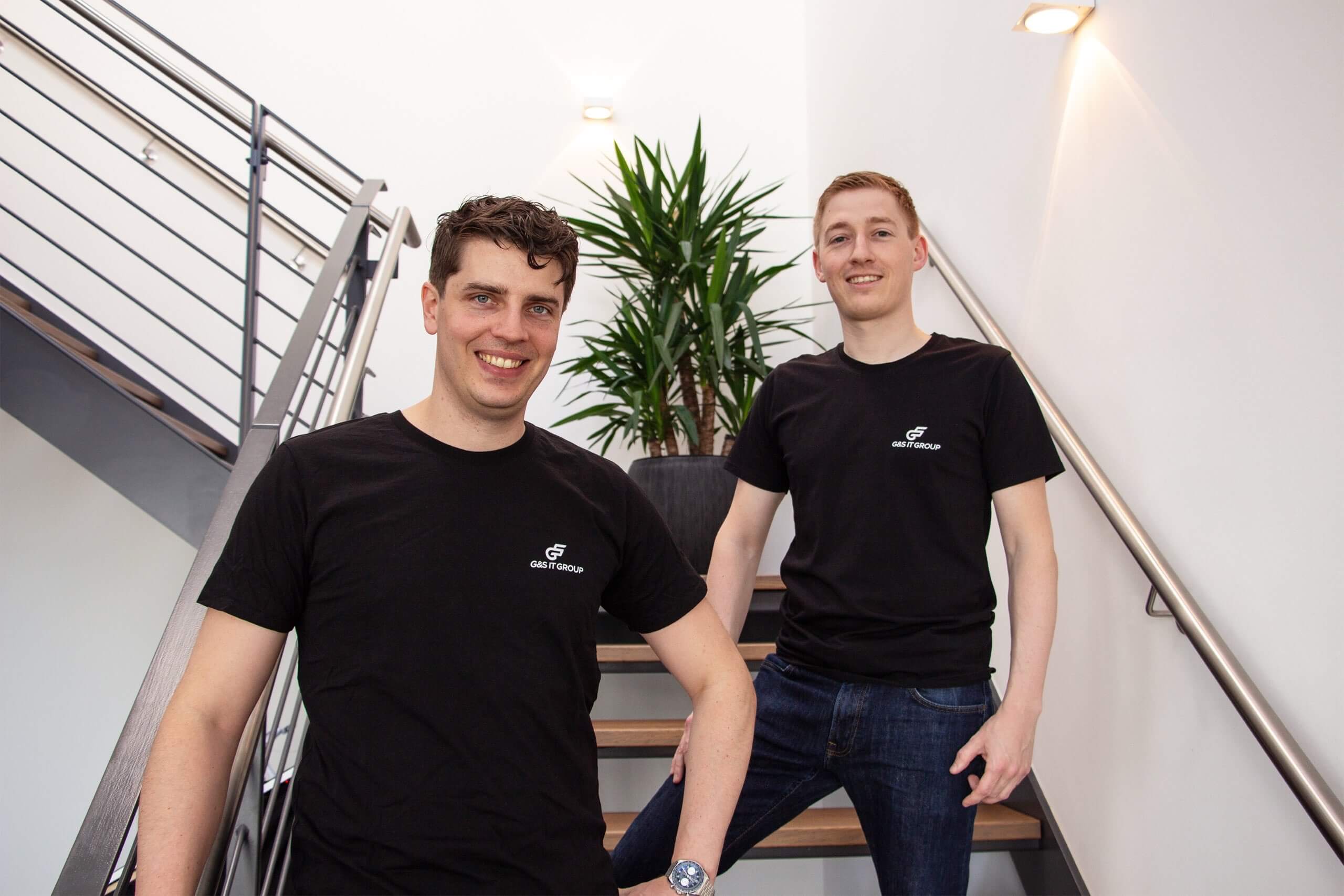 Zwei lächelnde Männer im schwarzen T-Shirt der G&S IT Group stehen gemeinsam auf einer modernen Holztreppe im Büro.