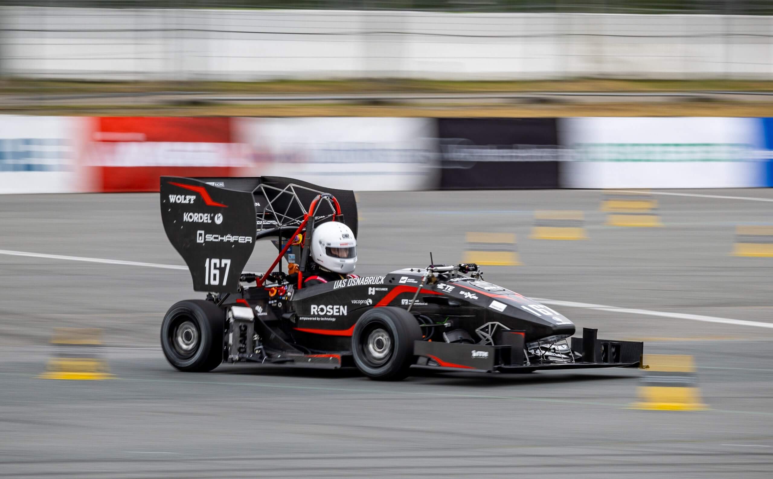Ein Rennfahrer mit weißem Helm fährt mit einem schwarzen Formel-Rennwagen dynamisch über eine Teststrecke.