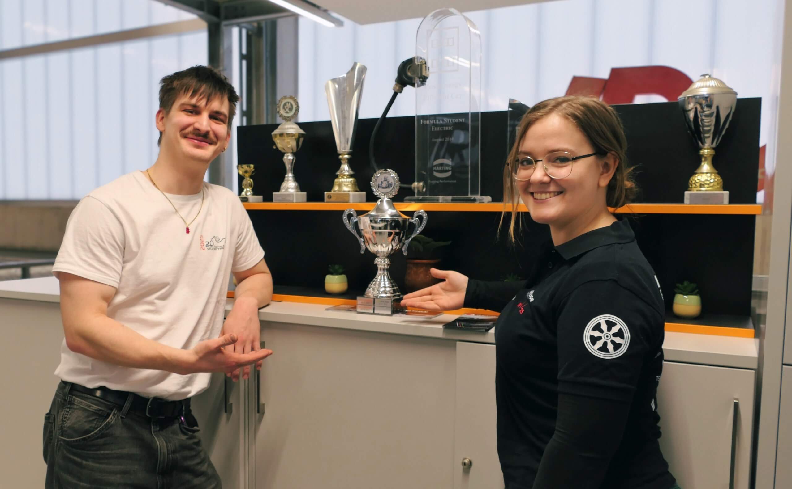 Zwei junge Menschen stehen lächelnd neben einem Regal voller Pokale und zeigen stolz auf eine Trophäe.