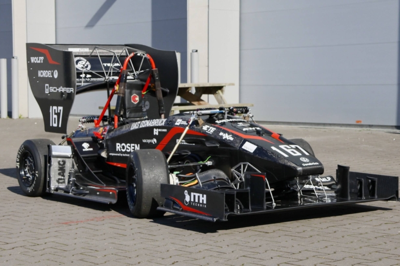 Ein kleiner, schwarz-roter Rennwagen mit großem Heckflügel steht schräg von vorne auf einem gepflasterten Platz.
