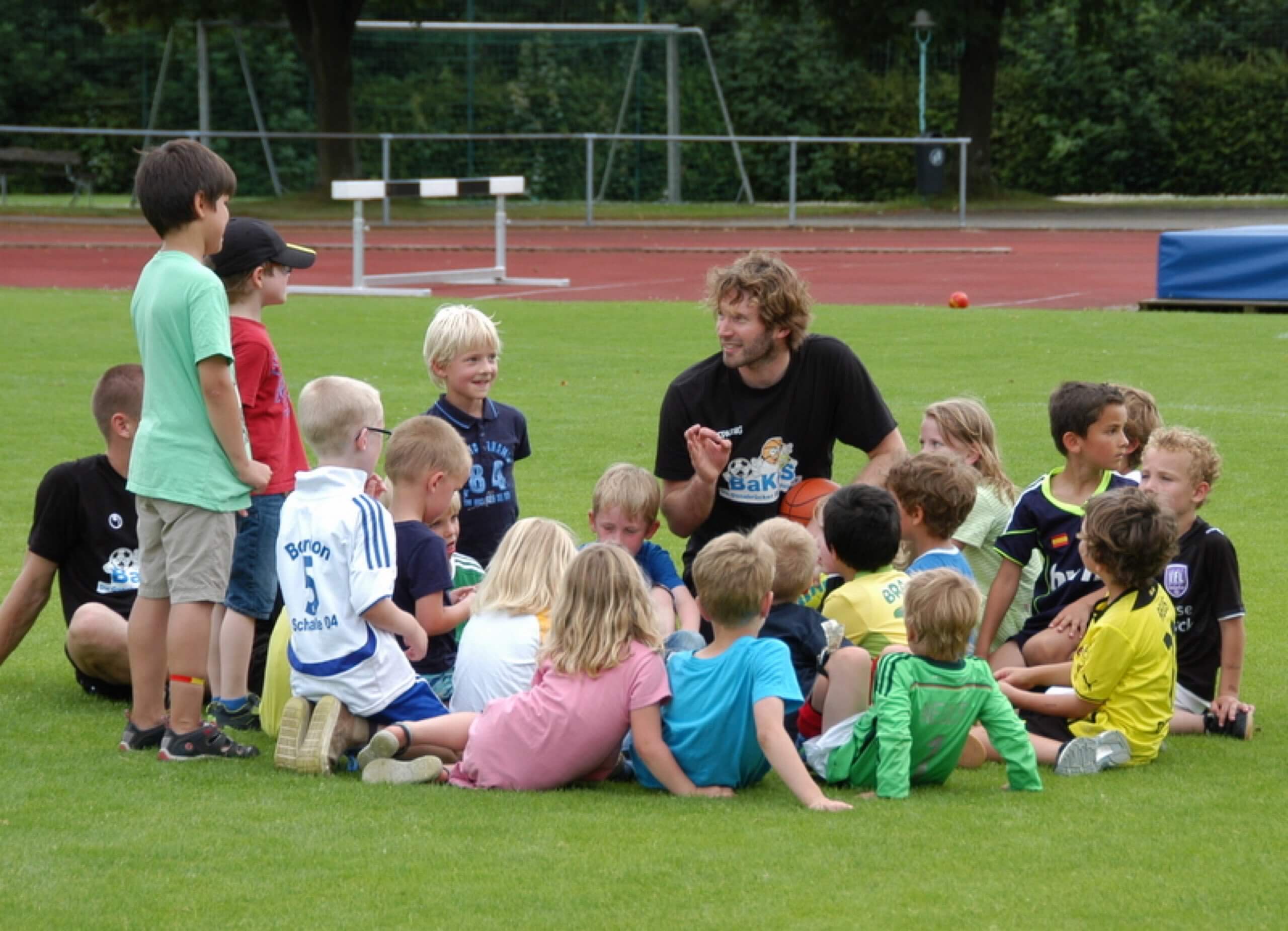 Ein Trainer sitzt mit einer Gruppe kleiner Kinder im Kreis auf einem Rasensportplatz und erklärt etwas.