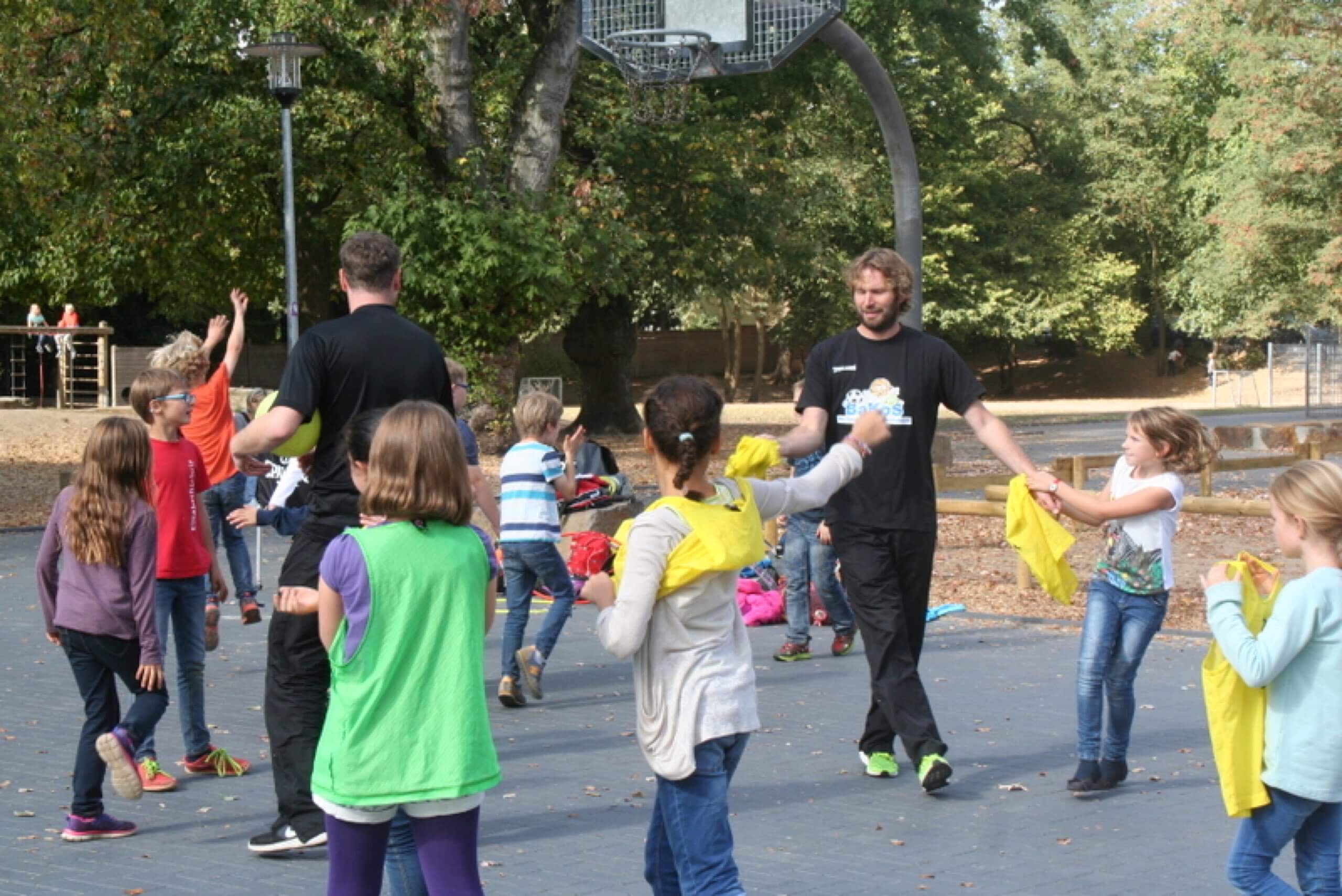 Ein Mann und mehrere Kinder spielen ein Bewegungsspiel mit gelben Tüchern auf einem Basketballplatz.