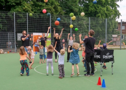 Kinder und Betreuer werfen auf einem Sportplatz gemeinsam bunte Bälle gleichzeitig hoch in die Luft.