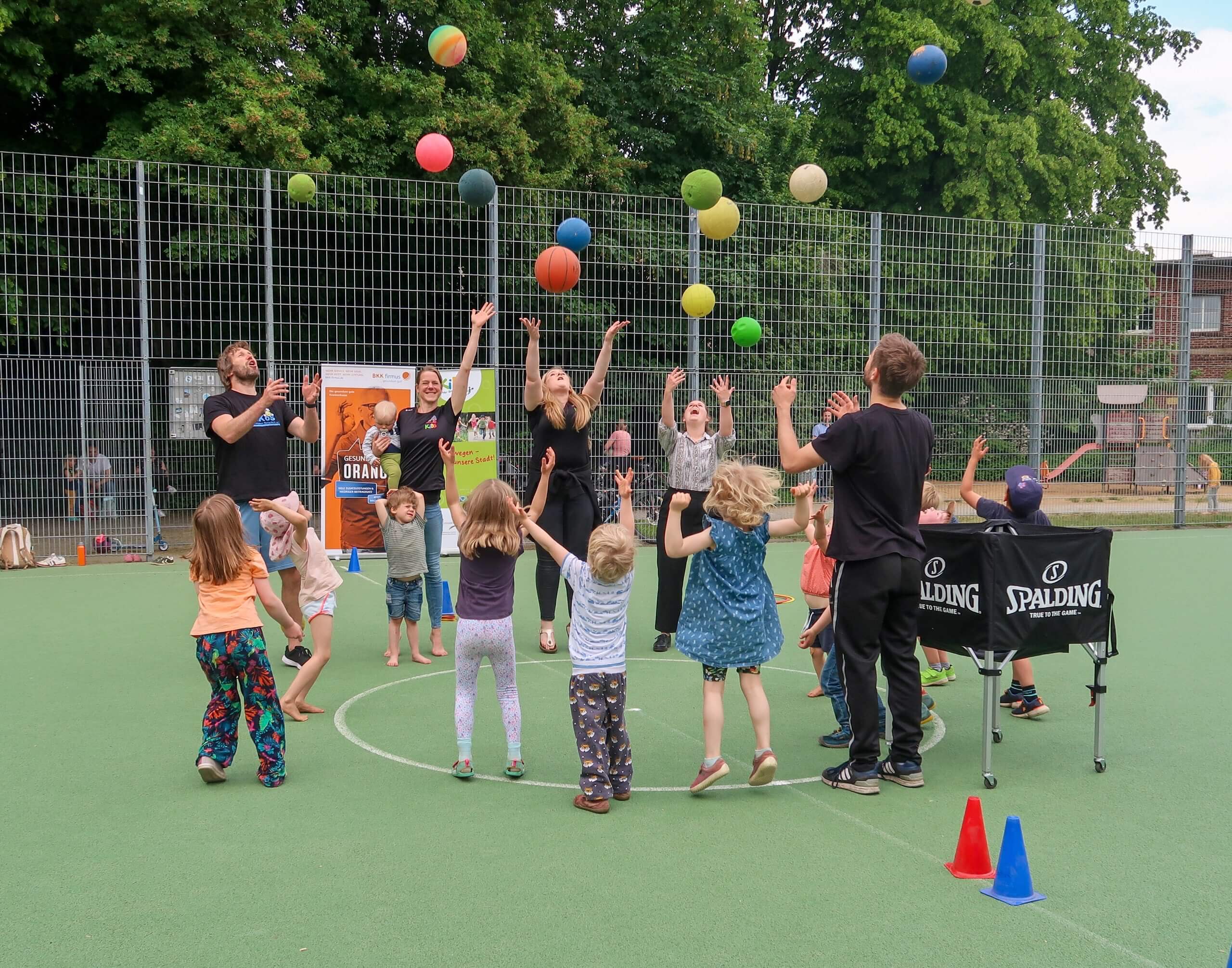 Kinder und Betreuer werfen auf einem Sportplatz gemeinsam bunte Bälle gleichzeitig hoch in die Luft.