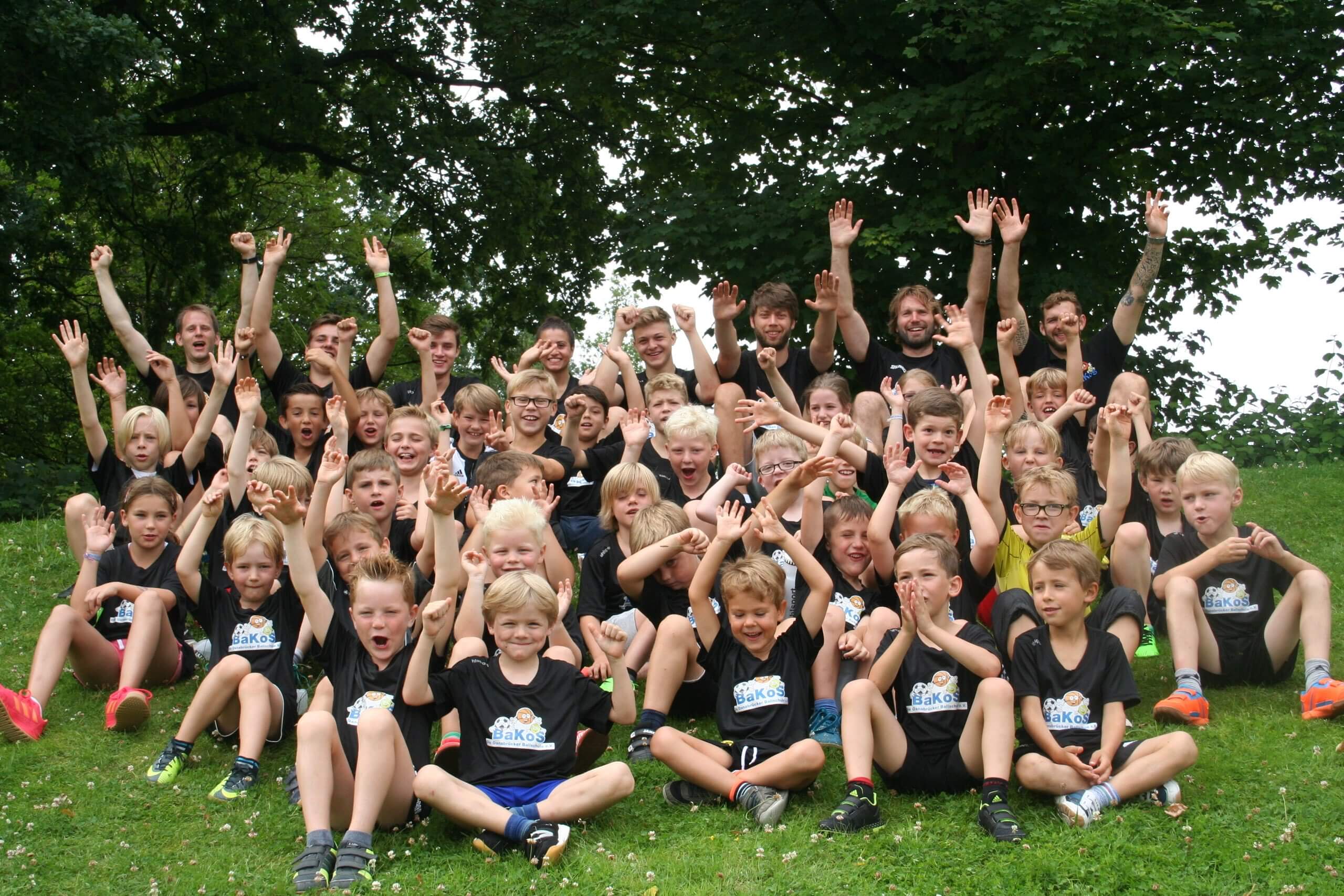Eine große Kindergruppe und ihre Betreuer sitzen im Gras und jubeln mit erhobenen Händen in die Kamera.
