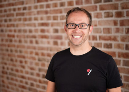 Ein lächelnder Mann mit Brille und schwarzem T-Shirt steht vor einer rot-braunen Backsteinwand.