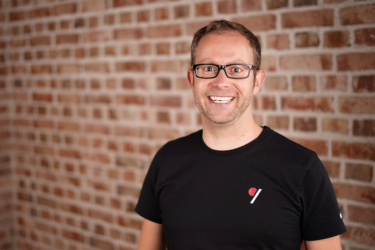 Ein lächelnder Mann mit Brille und schwarzem T-Shirt steht vor einer rot-braunen Backsteinwand.