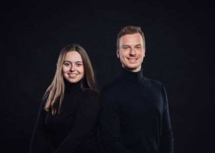 Porträtfoto einer jungen Frau und eines jungen Mannes, beide lächelnd in schwarzen Rollkragenpullovern vor schwarzem Hintergrund.