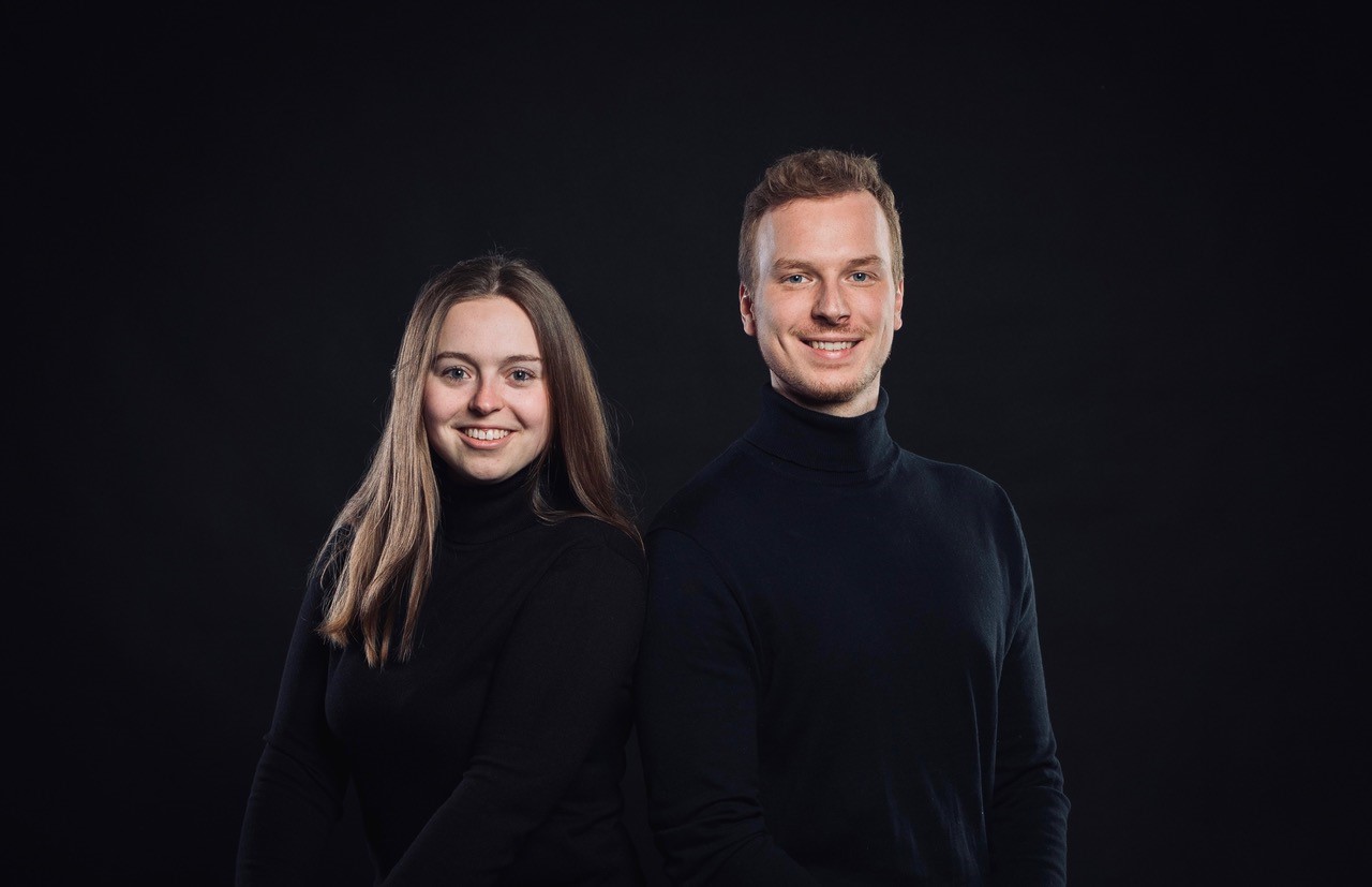 Porträtfoto einer jungen Frau und eines jungen Mannes, beide lächelnd in schwarzen Rollkragenpullovern vor schwarzem Hintergrund.