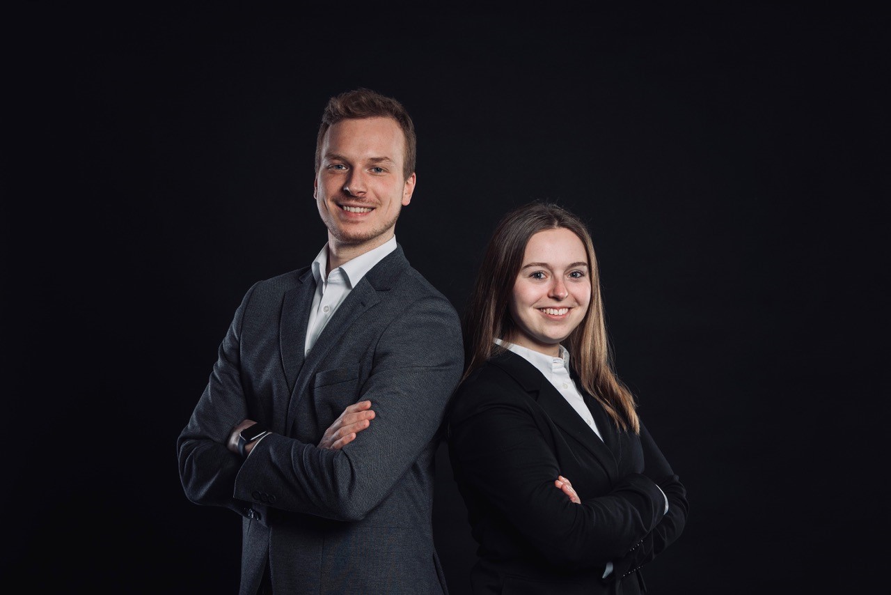 Porträtfoto eines Mannes in grauem Anzug und einer Frau in schwarzem Hosenanzug, beide lächelnd mit verschränkten Armen vor schwarzem Hintergrund.