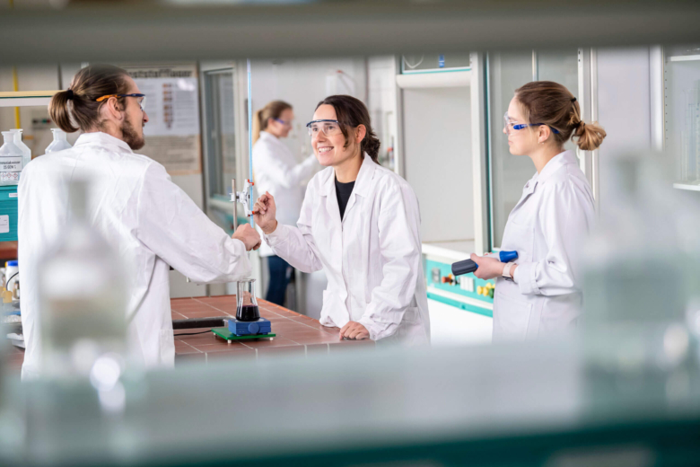 Drei Personen in weißen Laborkitteln und Schutzbrillen arbeiten zusammen an einem Chemie-Versuchsaufbau.