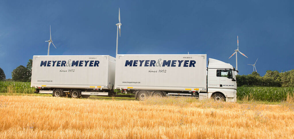 Ein weißer Lkw mit der Aufschrift „MEYER & MEYER“ steht auf einer Straße neben einem Getreidefeld mit Windkraftanlagen im Hintergrund.
