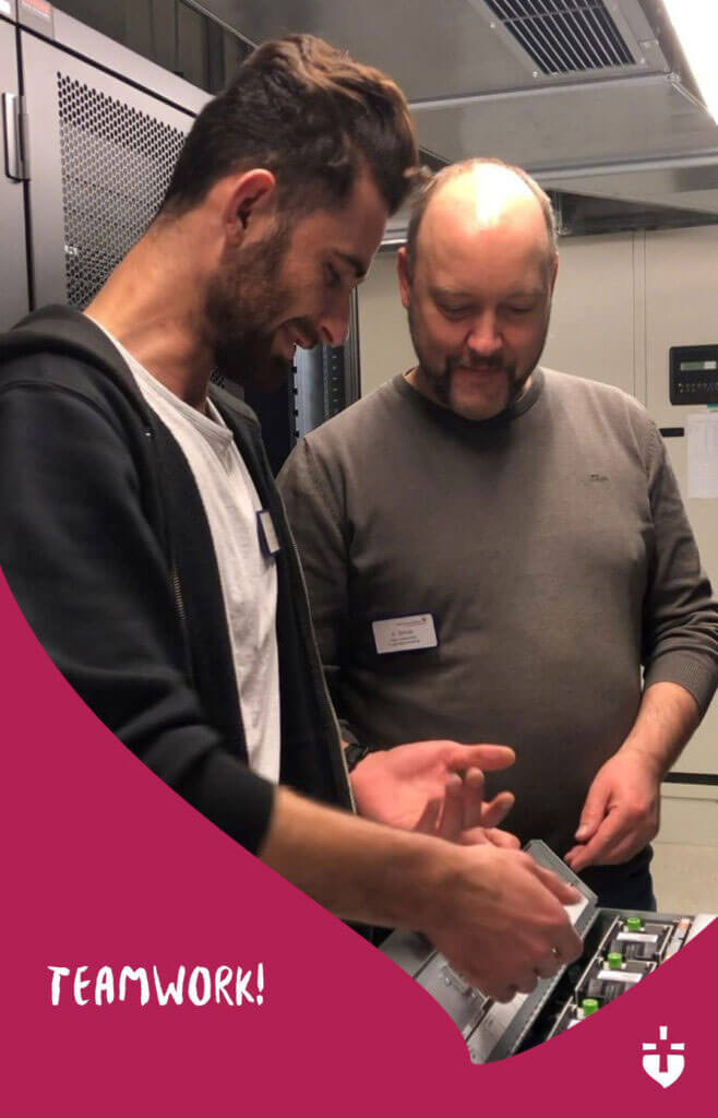 Absi Haj Hamdan und ein Kollege arbeiten gemeinsam an einer technischen Anlage. Im Vordergrund steht das Wort „TEAMWORK!“.