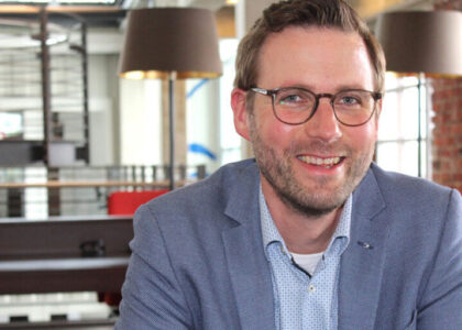 Porträtfoto von Adam Pietzka in einem modernen Büro. Er trägt eine Brille und ein blaues Sakko über einem hellblauen Hemd und lächelt. Der Hintergrund ist leicht unscharf.
