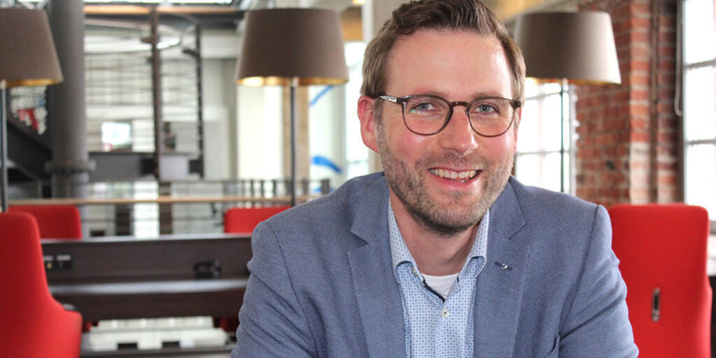Porträtfoto von Adam Pietzka in einem modernen Büro. Er trägt eine Brille und ein blaues Sakko über einem hellblauen Hemd und lächelt. Der Hintergrund ist leicht unscharf.