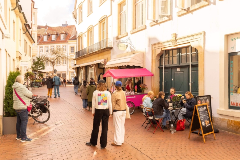Gäste sitzen vor einem historischen Gebäude an einem Straßencafé, während Passanten auf dem Platz flanieren und ein pinker Eisladenwagen steht.
