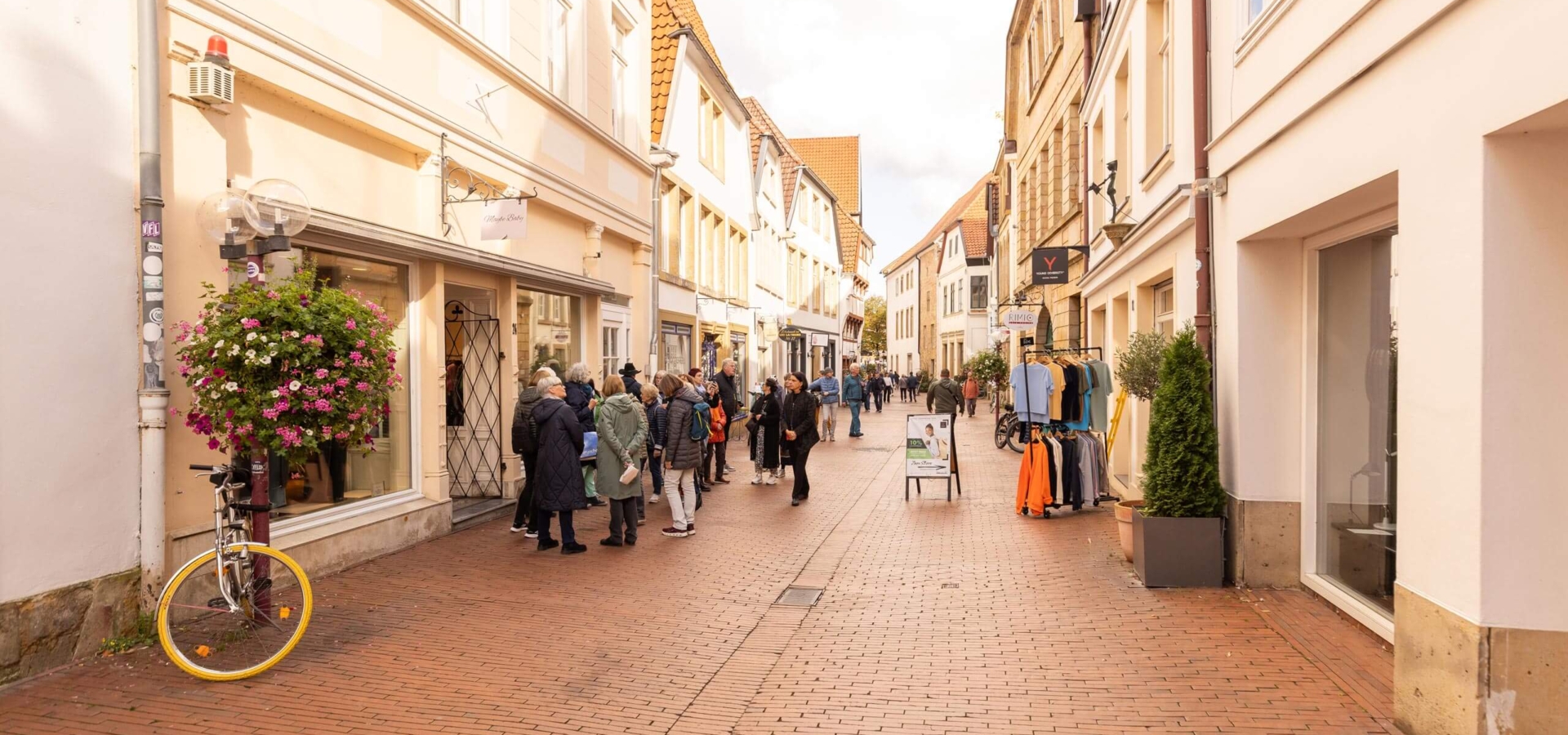 Fußgänger schlendern durch eine malerische Altstadtgasse mit historischen Fachwerkhäusern, Kopfsteinpflaster und blühenden Blumenampeln.