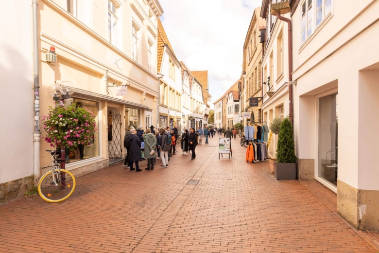 Fußgänger schlendern durch eine malerische Altstadtgasse mit historischen Fachwerkhäusern, Kopfsteinpflaster und blühenden Blumenampeln.