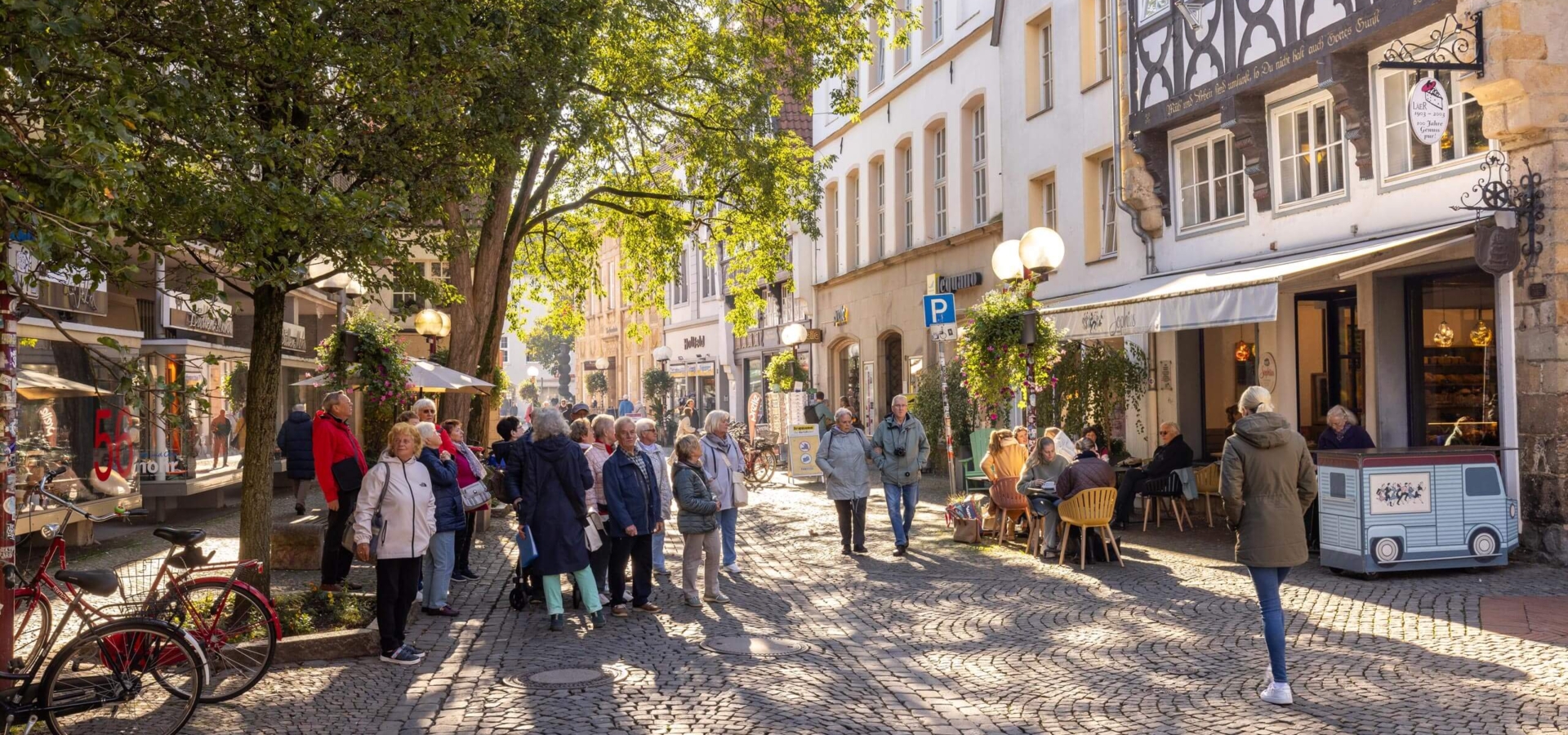 Zahlreiche Menschen gehen bei Sonnenschein über eine alte Kopfsteinpflasterstraße mit schattigen Bäumen und historischen Fachwerkhäusern.