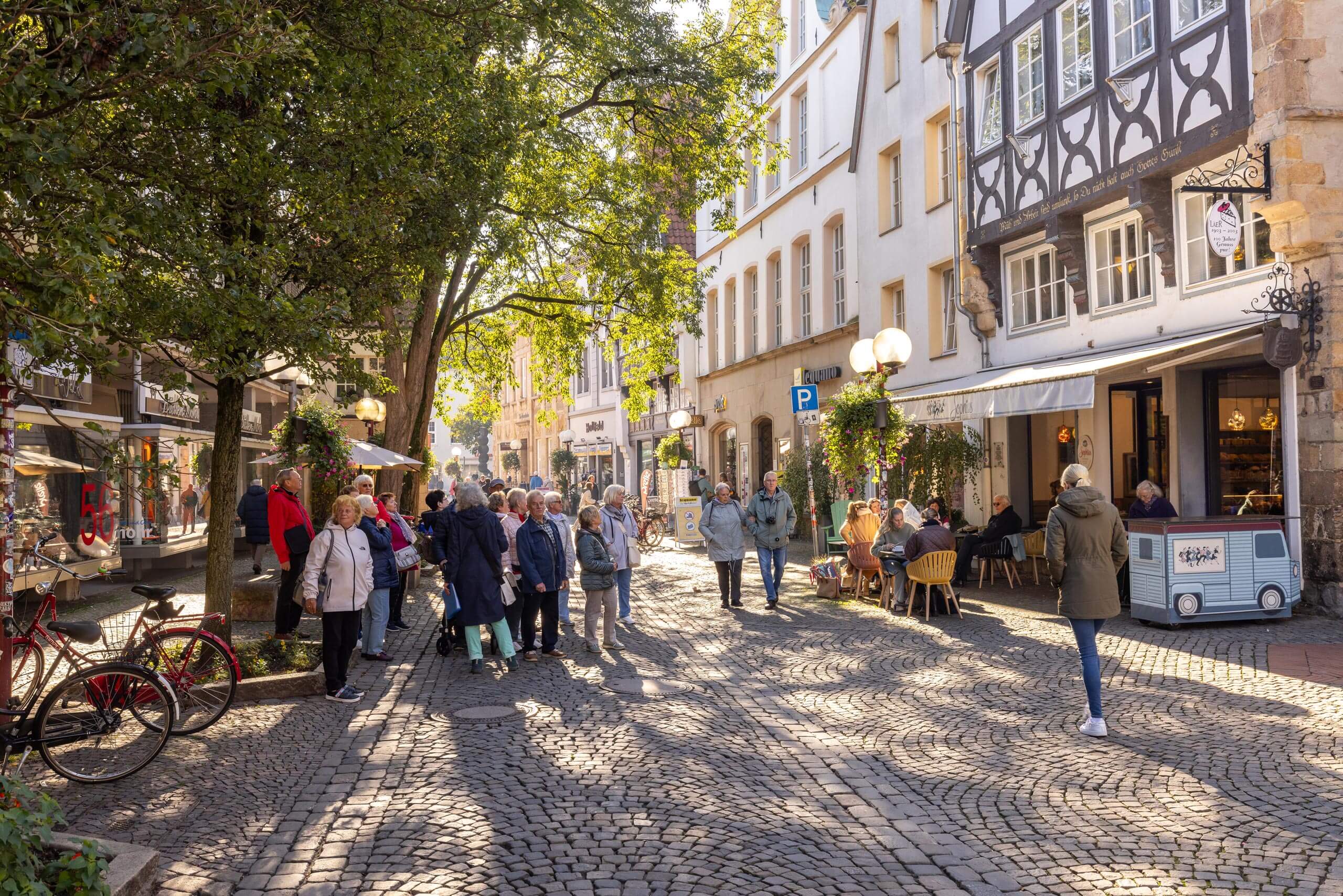 Zahlreiche Menschen gehen bei Sonnenschein über eine alte Kopfsteinpflasterstraße mit schattigen Bäumen und historischen Fachwerkhäusern.