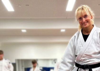 Andrea Goslar lächelt im weißen Judo-Anzug mit schwarzem Gürtel in einer Trainingshalle. Im verschwommenen Hintergrund sind weitere Personen im Judo-Anzug zu sehen.