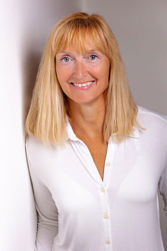 Porträt von Andrea Goslar mit blonden Haaren und weißer Bluse, die lächelnd an einer weißen Wand lehnt.