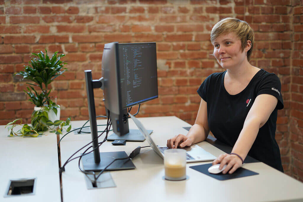 Andrea Suckro sitzt lächelnd an einem Schreibtisch vor zwei Monitoren und arbeitet am Laptop.