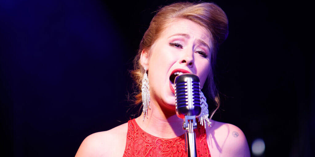 Anne Bischow singt leidenschaftlich im roten Kleid in ein klassisches Retro-Mikrofon auf einer Bühne vor dunklem, blau beleuchtetem Hintergrund.