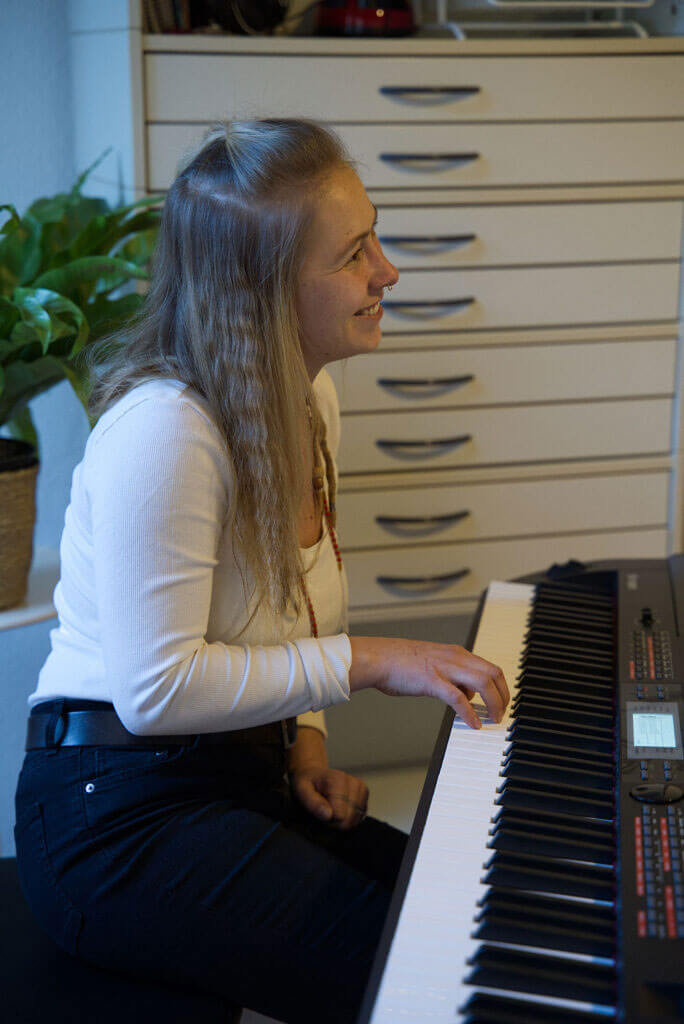 Anne Bischow sitzt lächelnd im Profil an einem E-Piano und spielt mit der rechten Hand auf den Tasten.