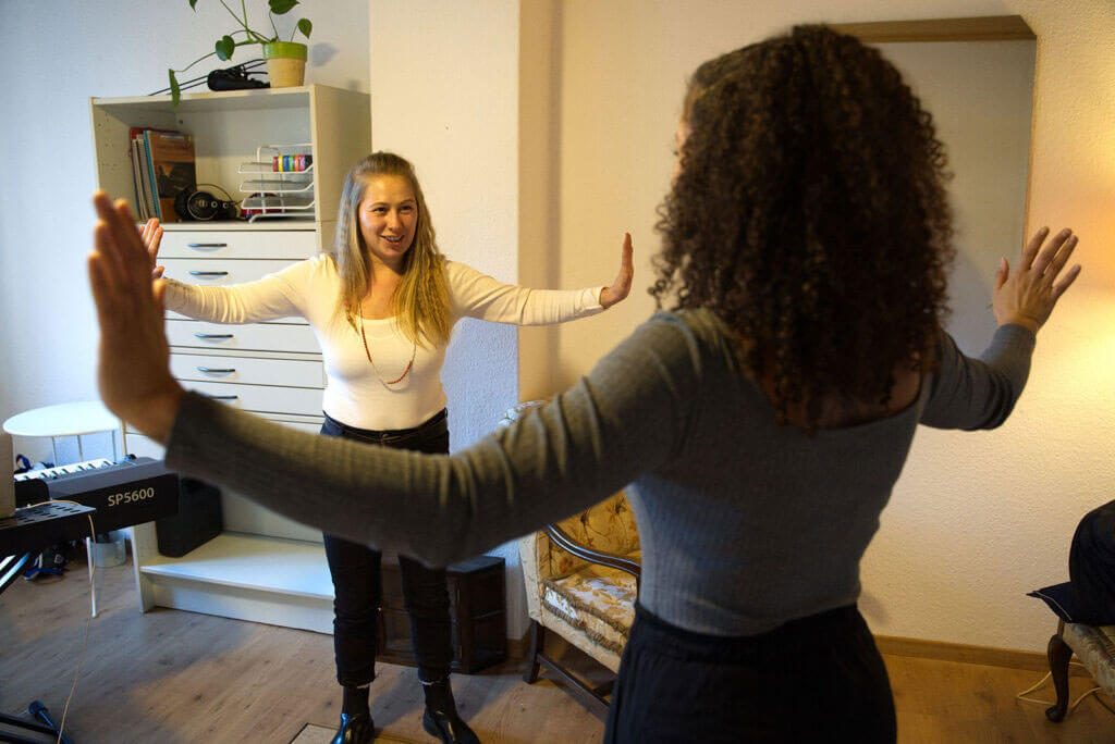 Anne Bischow macht in einem Raum eine gemeinsame Atem- oder Stimmübung mit einer Teilnehmerin, beide mit weit ausgestreckten Armen.