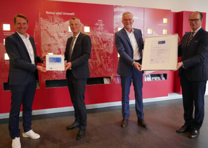 Vier Männer in Anzügen halten gemeinsam zwei gerahmte Urkunden vor einer roten Wandtafel in einem Ausstellungsraum.