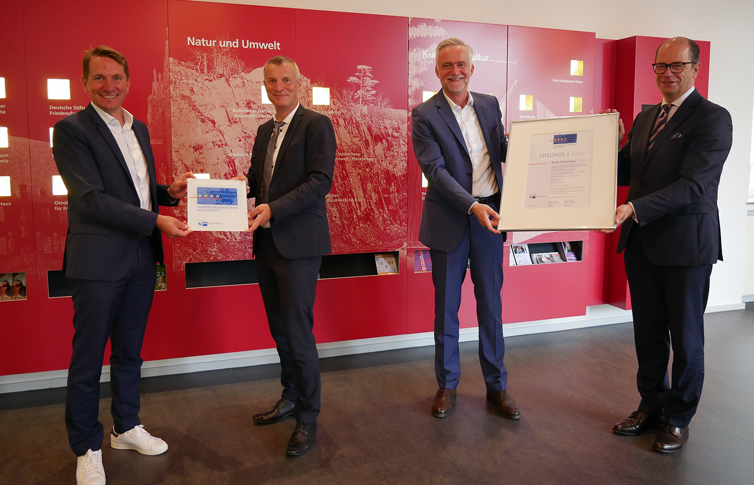 Vier Männer in Anzügen halten gemeinsam zwei gerahmte Urkunden vor einer roten Wandtafel in einem Ausstellungsraum.