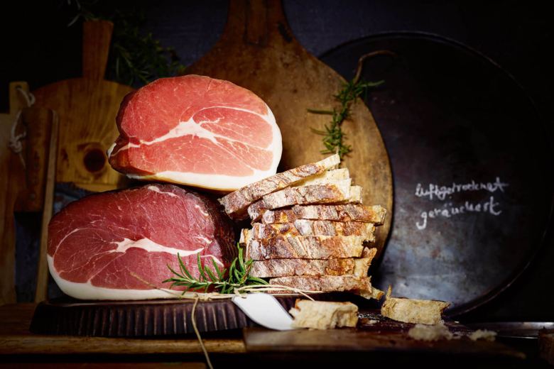 Zwei rohe Schinkenstücke, Brotscheiben und Rosmarin auf einem dunklen Holztisch vor rustikalen Holzbrettern und einer Metallplatte.