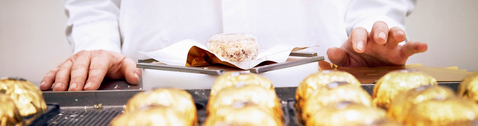 Blick über eine Reihe in Goldfolie gewickelter Kugeln auf eine Person in weißer Arbeitskleidung, die eine Kugel auf Papier platziert.