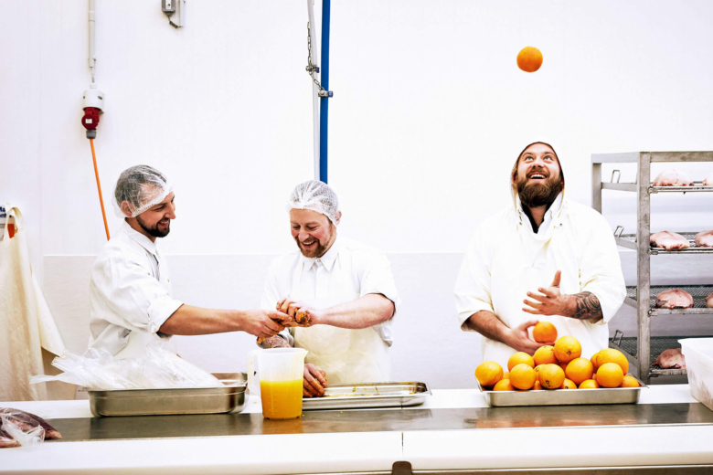 Drei lächelnde Männer in weißer Arbeitskleidung und Haarnetzen in einer Großküche; einer presst Orangen, einer jongliert mit einer Orange.