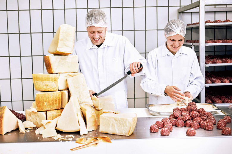 Zwei lächelnde Personen in weißer Arbeitskleidung verarbeiten Fleischbällchen und schneiden Stücke von einem großen Stapel Parmesanrädern ab.
