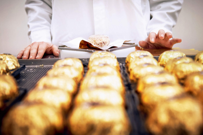 Person in weißer Arbeitskleidung bei der Herstellung von Saltufo-Spezialitäten. Im Vordergrund stehen in Goldfolie gewickelte Kugeln.