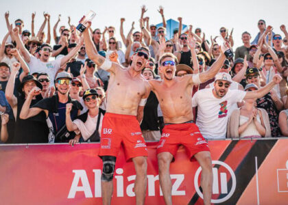 Beachvolleyballer Bennet Poniewaz und sein Teamkollege ohne Oberkörper in roten Shorts jubeln mit erhobenen Armen und Trophäen vor einer großen, klatschenden Zuschauermenge bei der German Beach Tour.